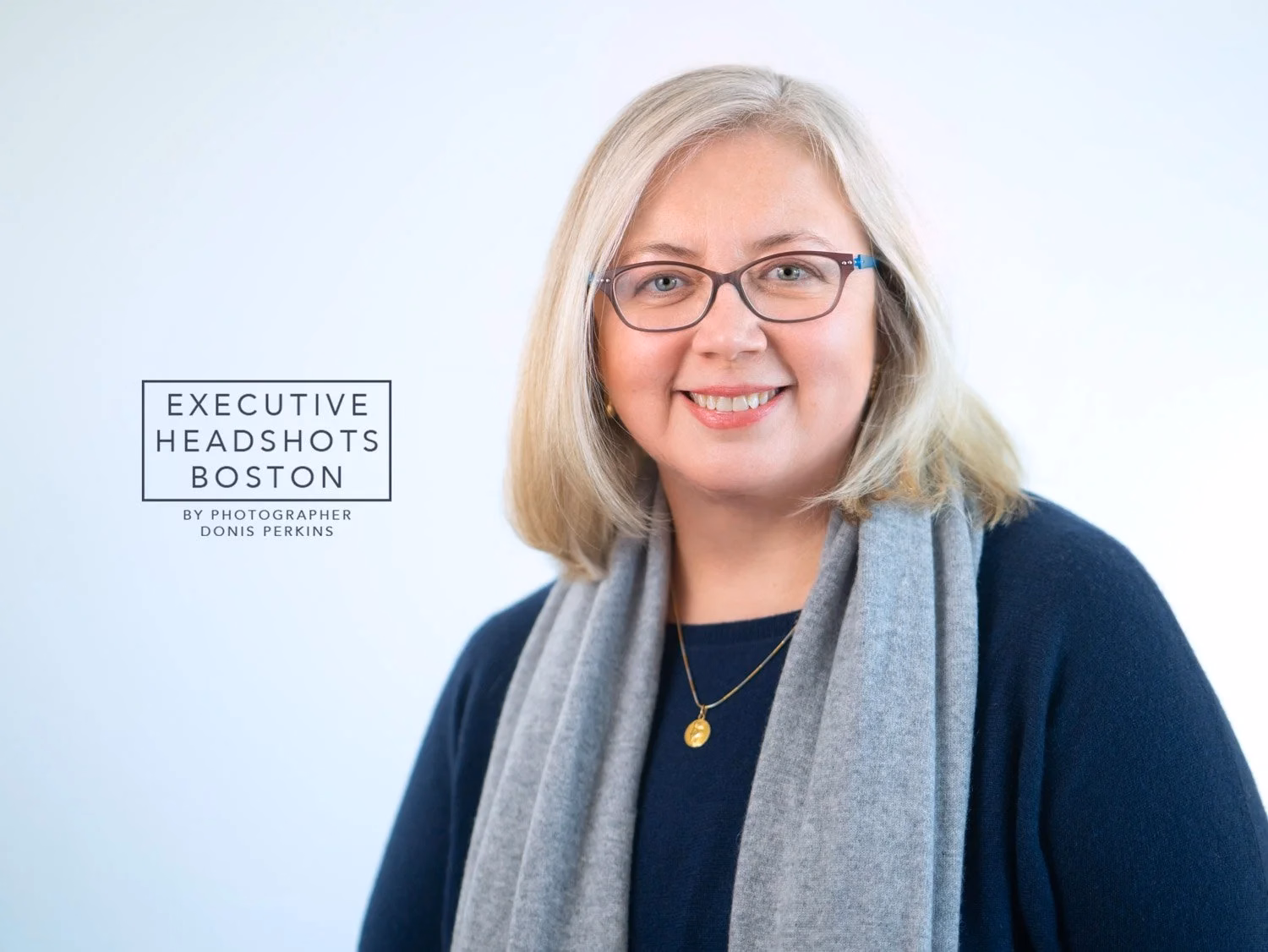 A smiling woman with shoulder-length blonde hair and burgundy glasses in a navy sweater with a grey scarf and gold pendant, against a pale background