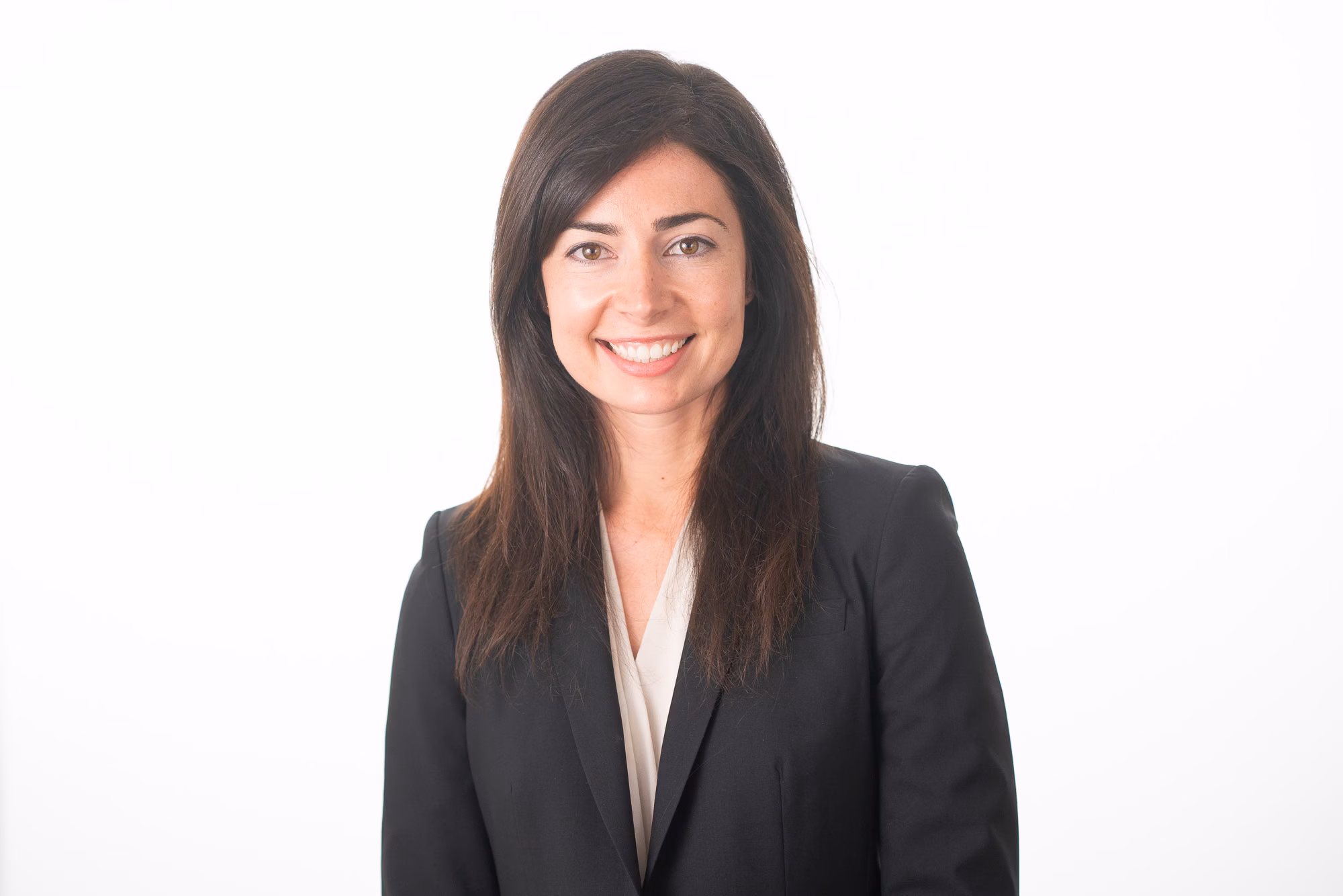 Woman with long hair wears a black blazer over a cream top, smiling against a plain white backdrop