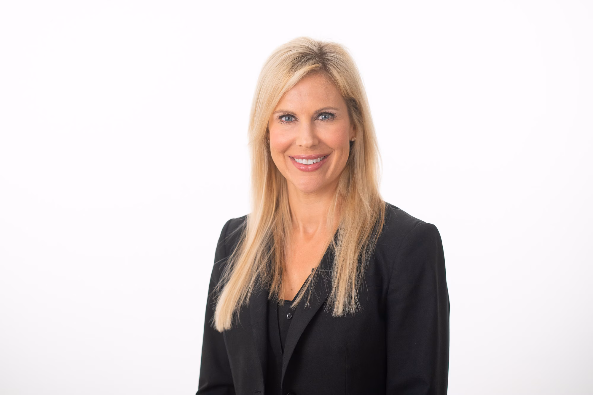 Long hair falls past the shoulders of a woman in a black blazer, smiling against a plain white backdrop