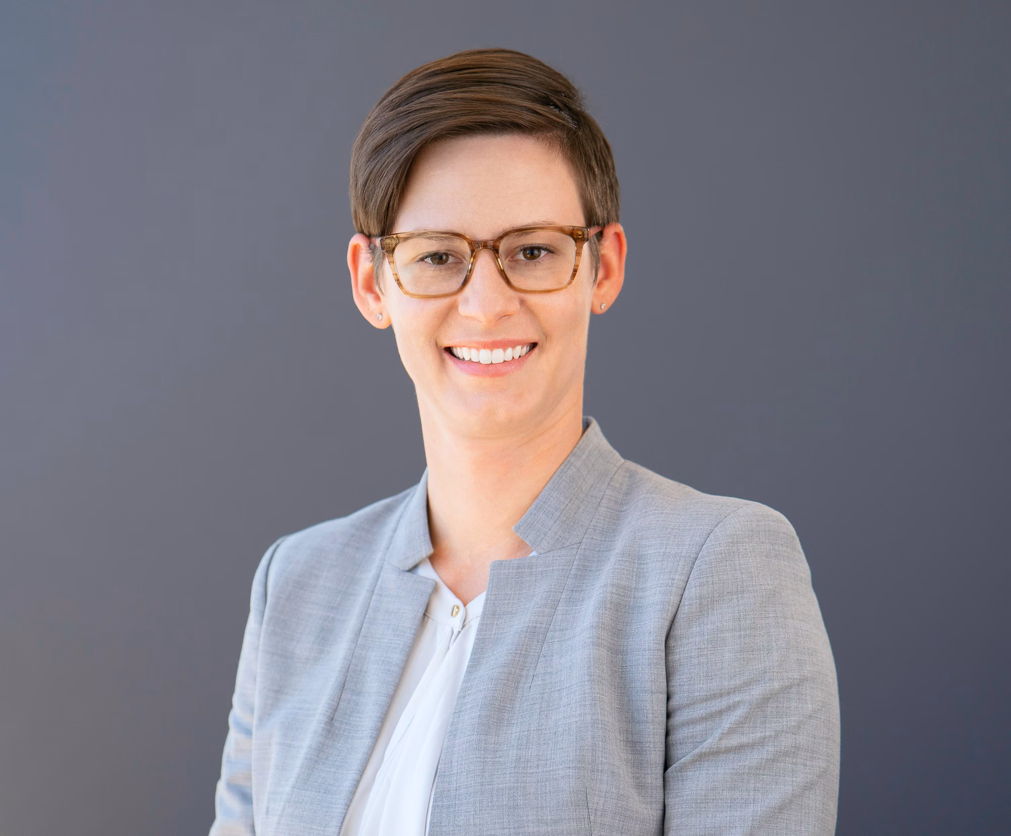 Wearing a light grey blazer over a white top, she smiles with tortoiseshell glasses against a plain grey backdrop