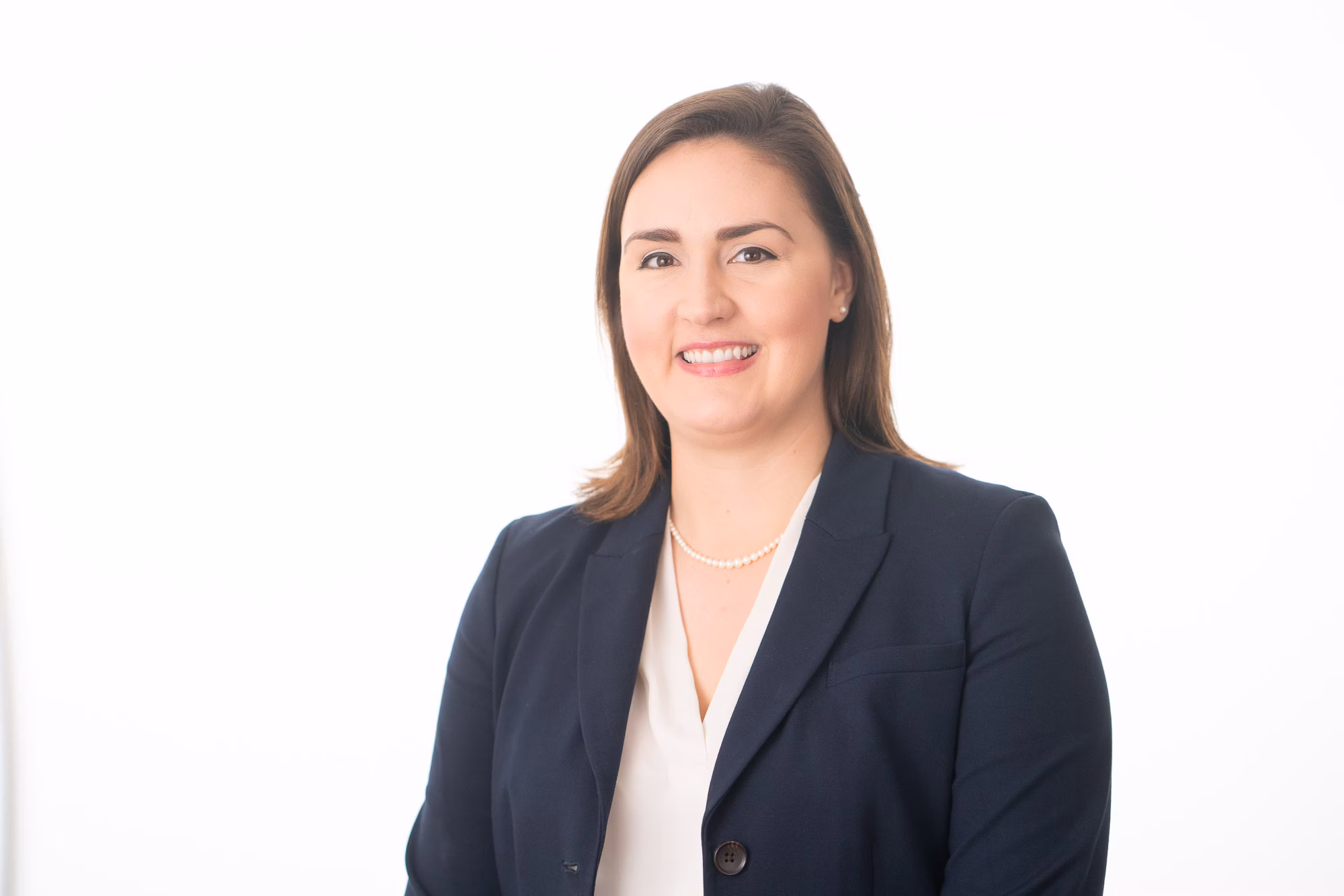 Woman in a tailored navy blazer and pearl necklace over a cream blouse, smiling against a plain white backdrop.