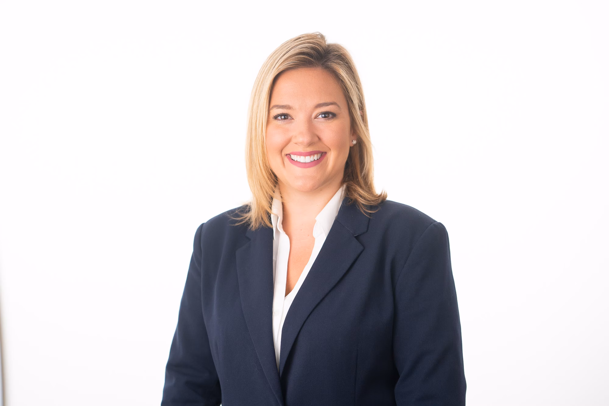 A woman in a navy blazer and white blouse smiles broadly against a plain white studio backdrop.