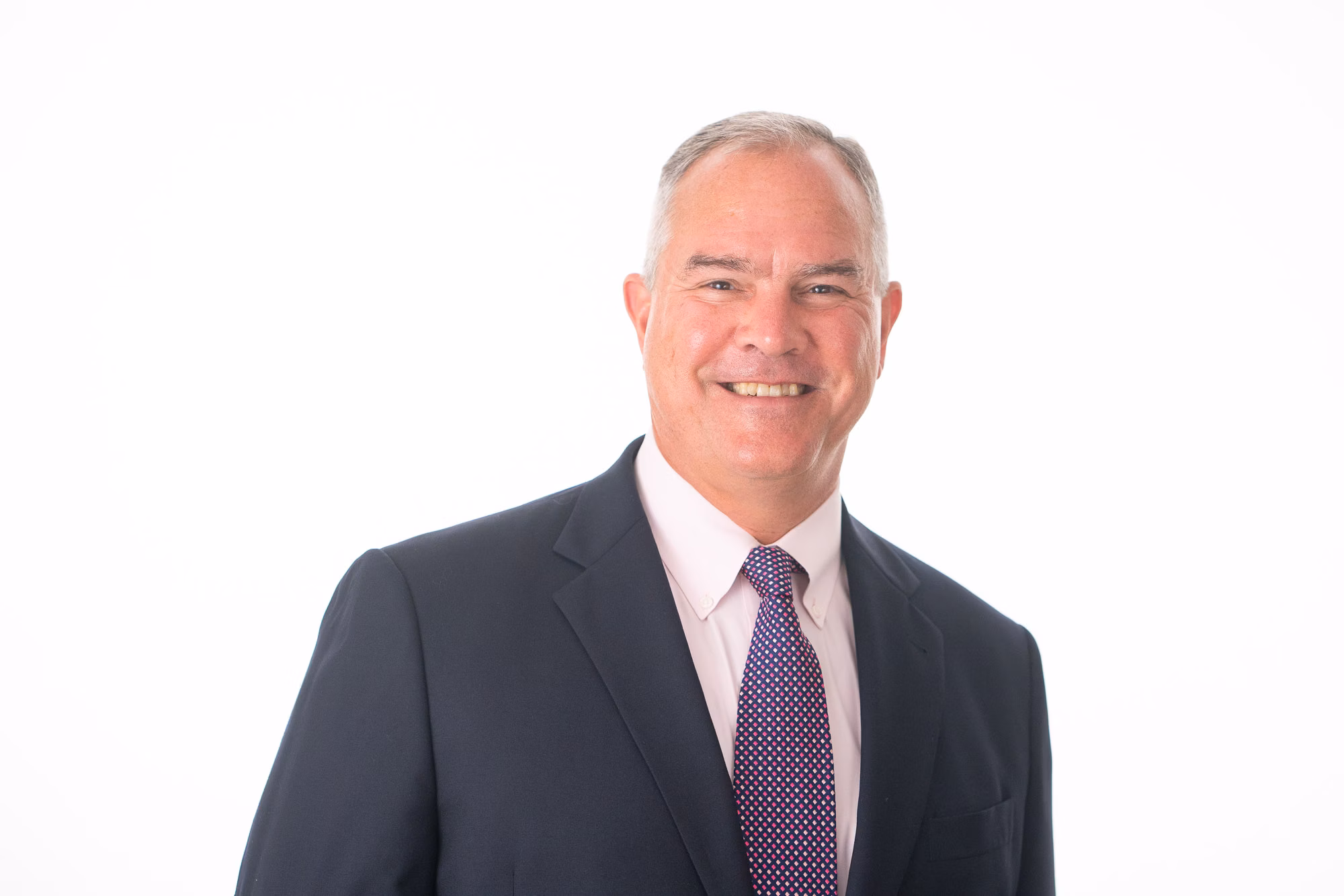 Man in a navy suit and pink shirt with a purple polka-dot tie smiles against a plain white background
