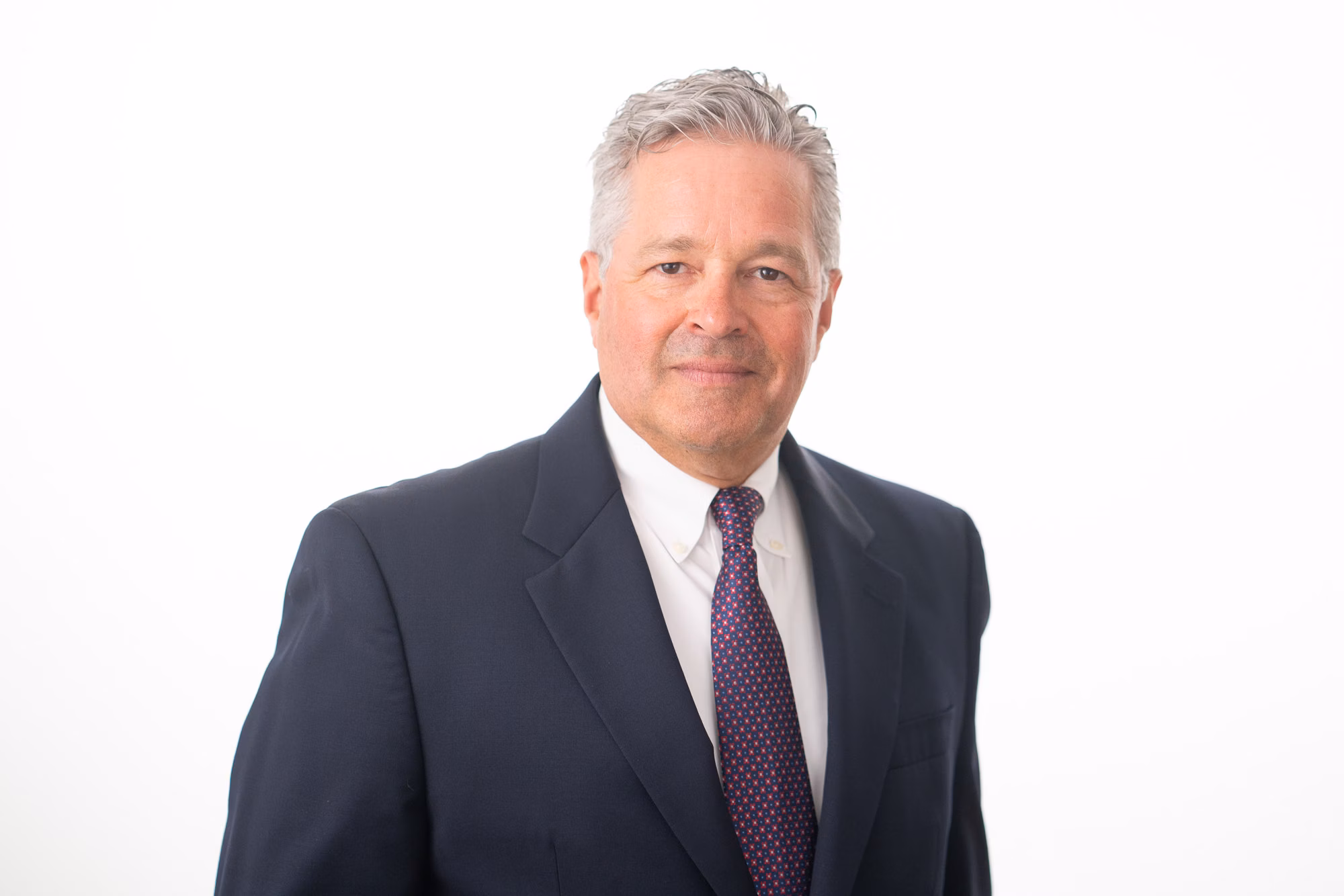Against a plain white backdrop, a man in a navy suit and dotted purple tie gives a relaxed, direct gaze.