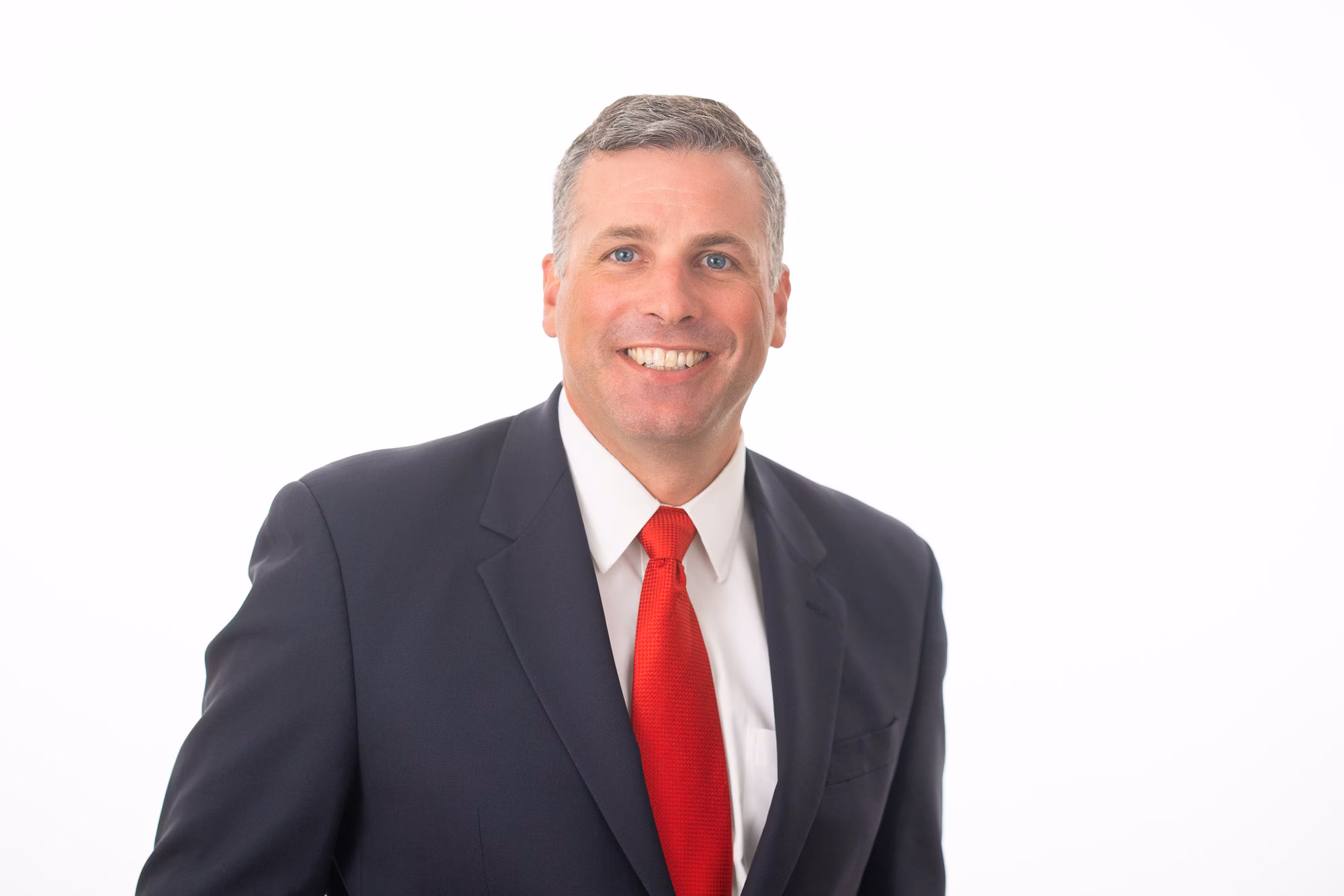 Wearing a navy suit with a bright red tie, he smiles broadly against a plain white background.
