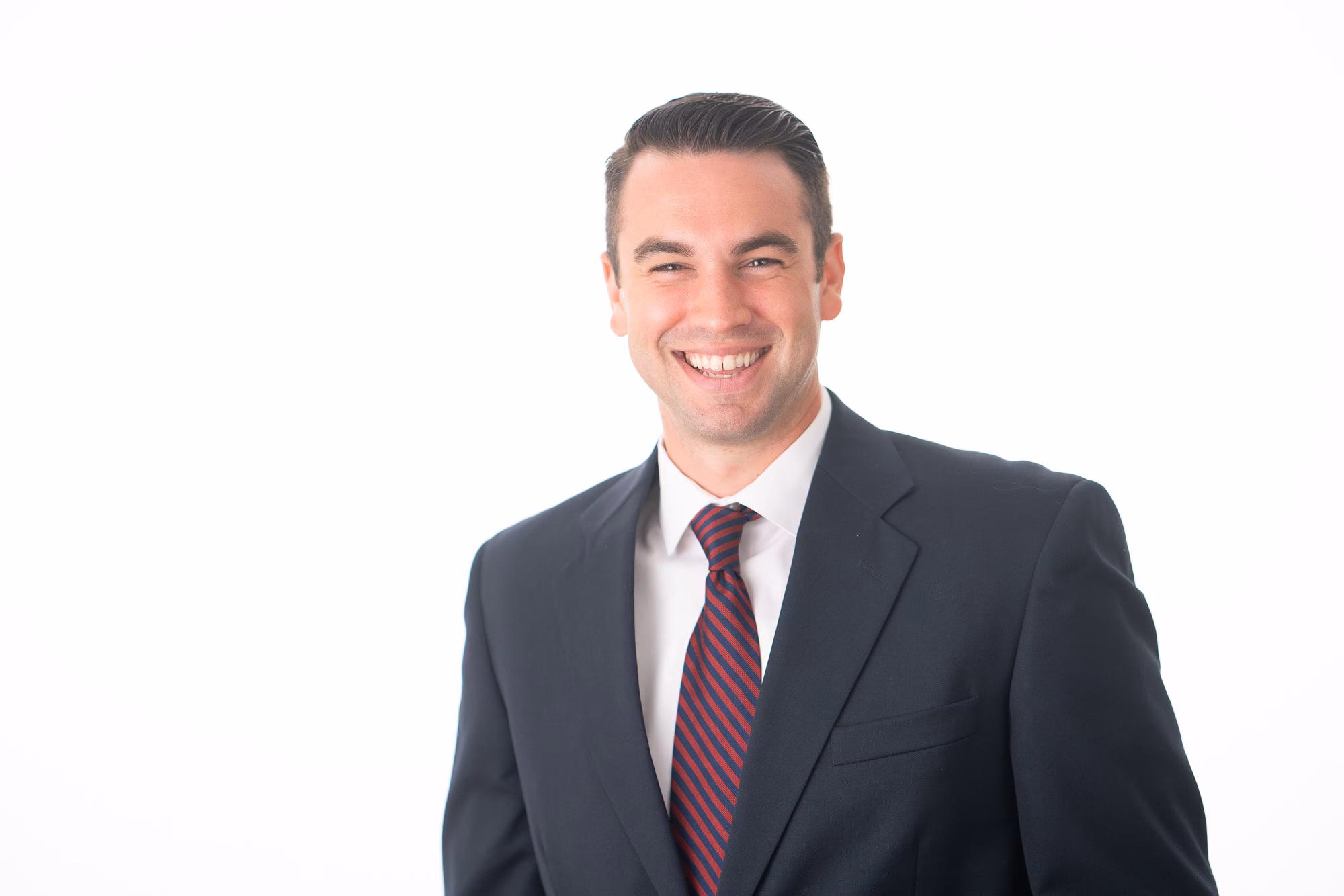 Man in a navy suit, maroon striped tie and crisp white shirt, grinning broadly against a plain white backdrop.