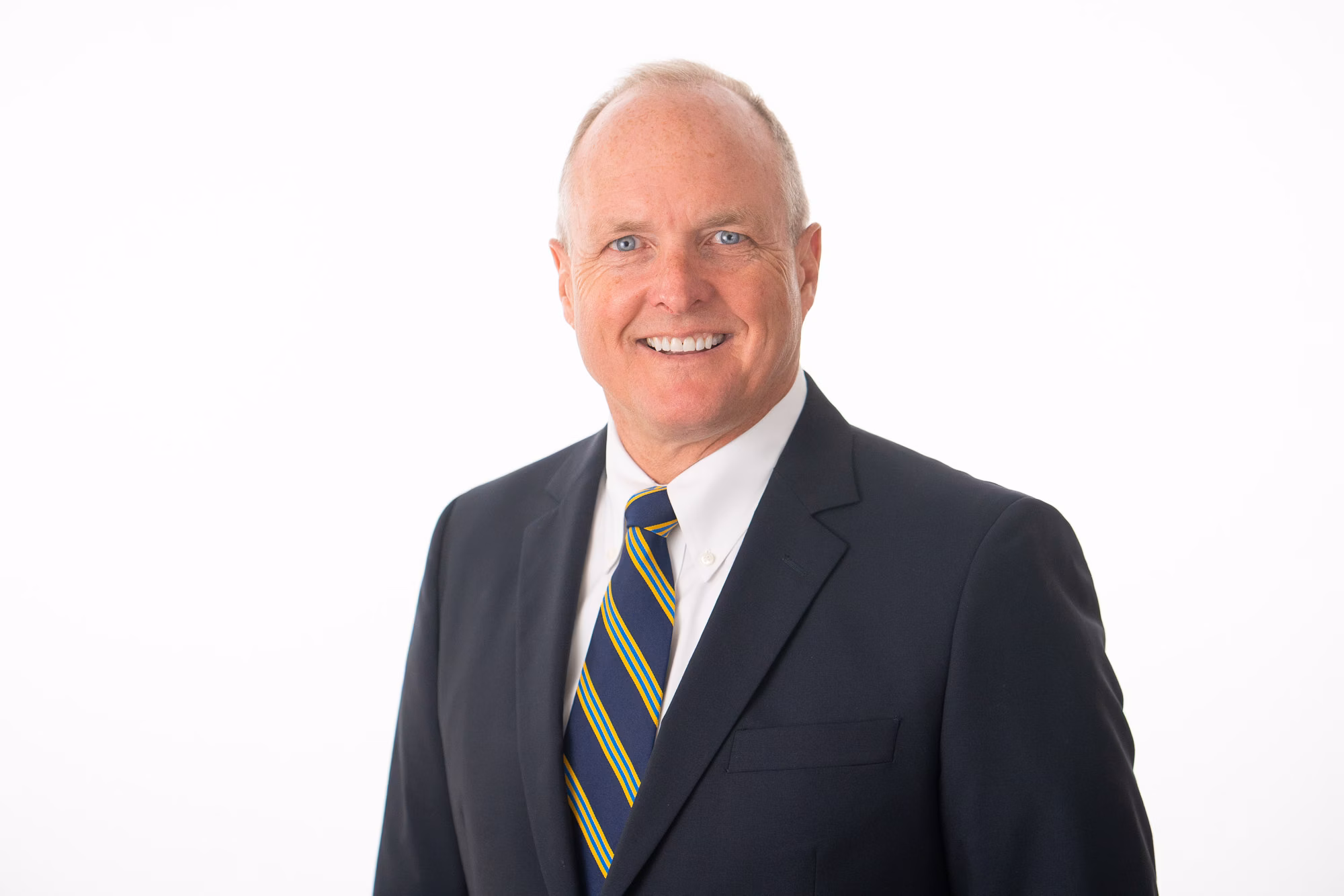 Man in a dark navy suit and blue-and-yellow striped tie smiles against a plain white backdrop