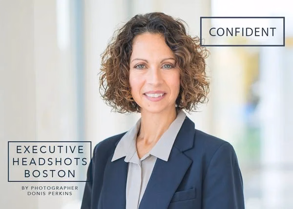 A smiling woman with shoulder-length curly brown hair in a navy blazer over a grey blouse, in a bright airy interior