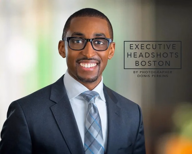 A smiling man with a short beard and black-framed glasses in a charcoal suit and pale blue plaid tie, against a blurred green and grey background