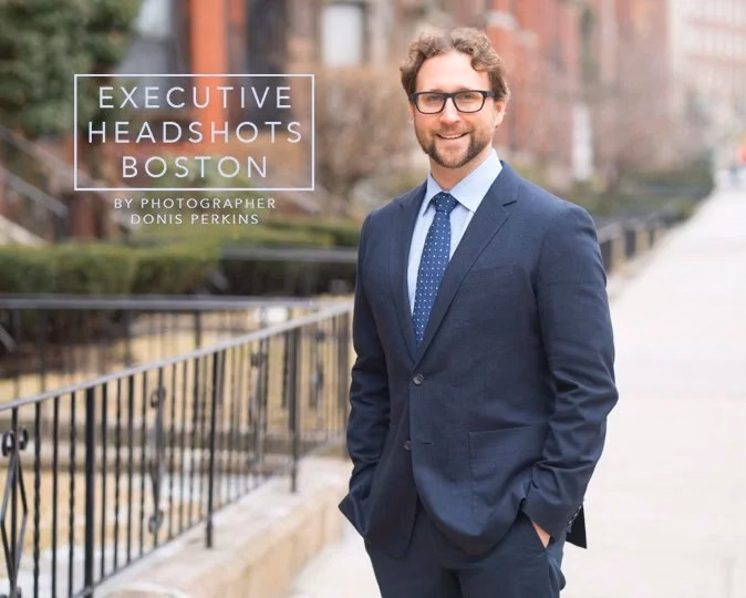 A bearded man in a navy suit and blue dotted tie standing on a Boston brownstone sidewalk with his hands in his pockets, an iron railing behind him