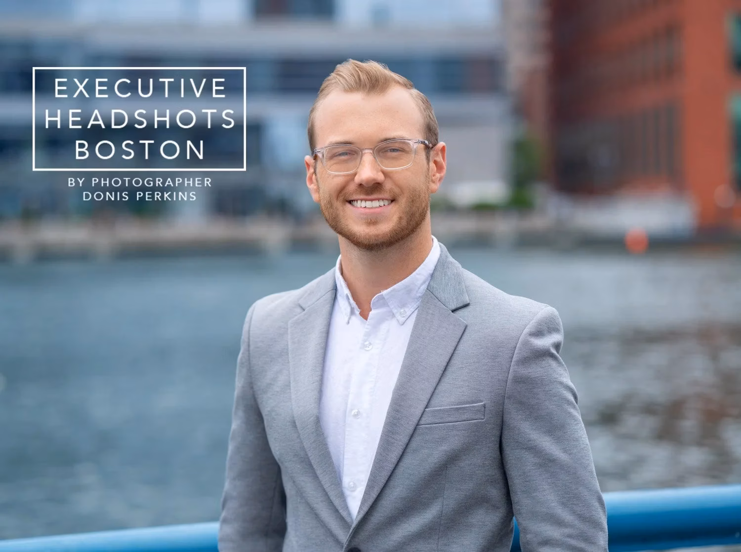 Smiling man with a short beard and clear-framed glasses in a light grey blazer beside Boston harbour water