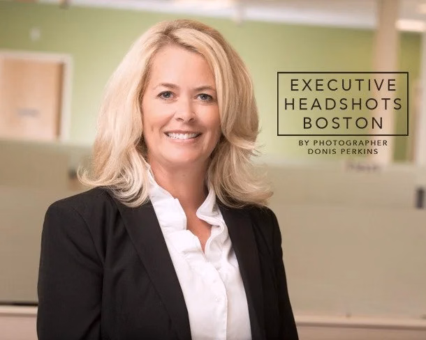 A smiling woman with shoulder-length blonde hair in a black blazer over a white ruffled blouse, in a green-walled office