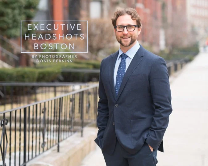 Smiling man with a beard and glasses in a navy suit and blue tie, hands in pockets, on a Boston brownstone sidewalk