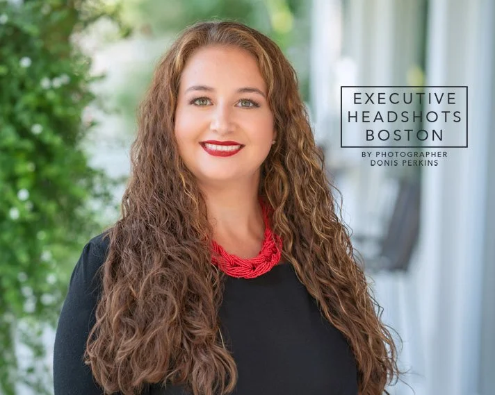 Smiling woman with long curly brown hair in a black top and red beaded necklace on a leafy porch
