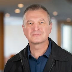 Donis Perkins, smiling slightly, with short grey hair, wearing a black jacket over a navy shirt against a softly blurred office background