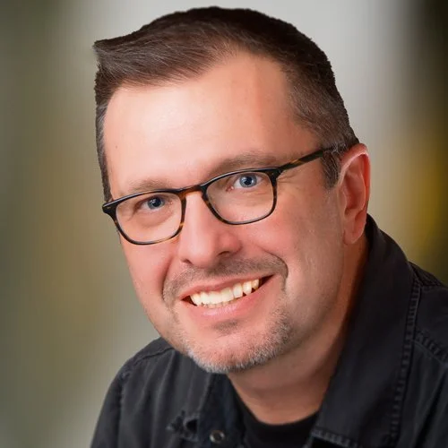 Smiling man with glasses and short greying hair in a dark shirt