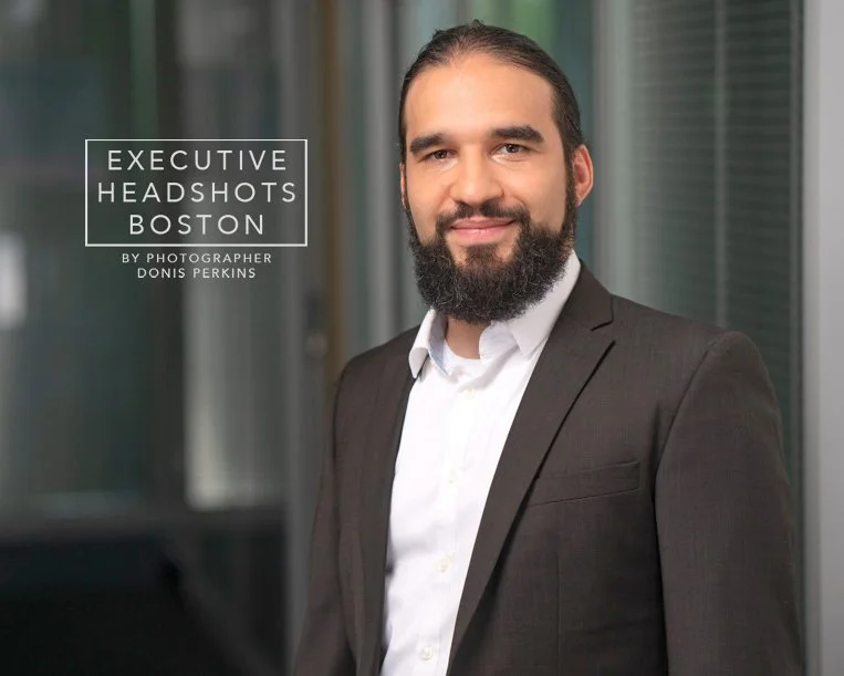 Smiling man with a full dark beard in a dark brown blazer over a white shirt in a glass-walled office