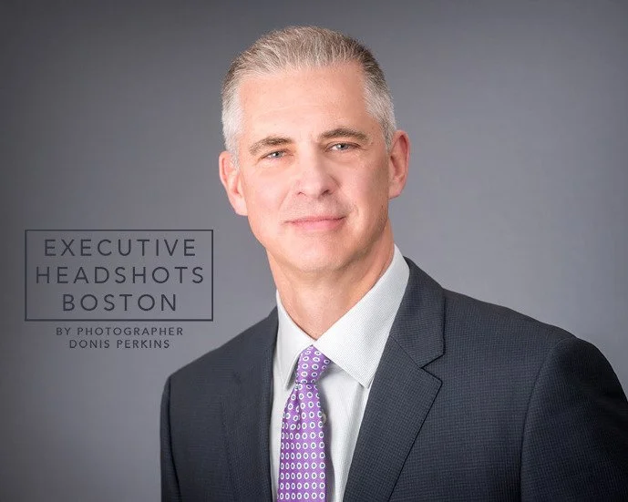 Man with short grey hair in a charcoal suit and purple patterned tie on a grey background