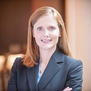Marsha Butler Gordon, smiling, with shoulder-length auburn hair, in a charcoal blazer over a blue top, arms folded