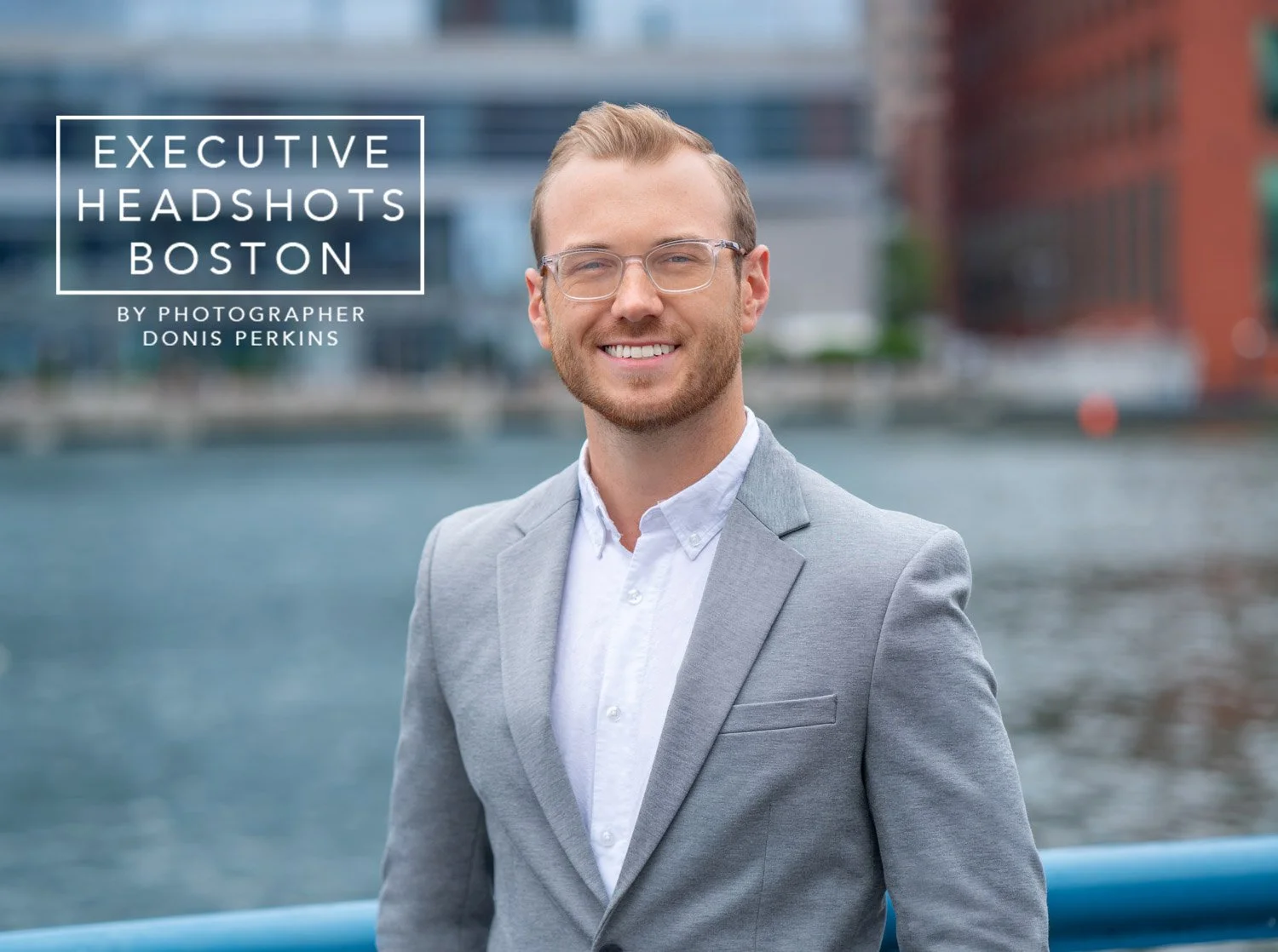 Smiling man with a short beard and clear-framed glasses in a light grey blazer beside Boston harbour water