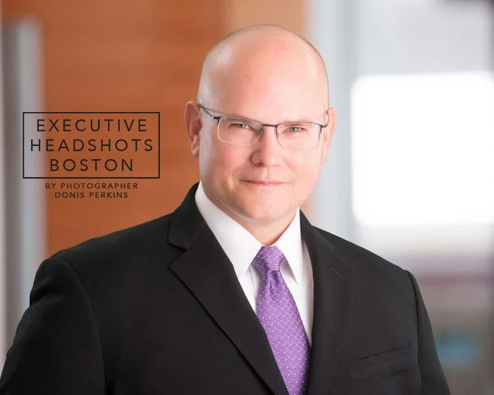 Bald man with glasses in a black suit, white shirt and purple tie in a bright office