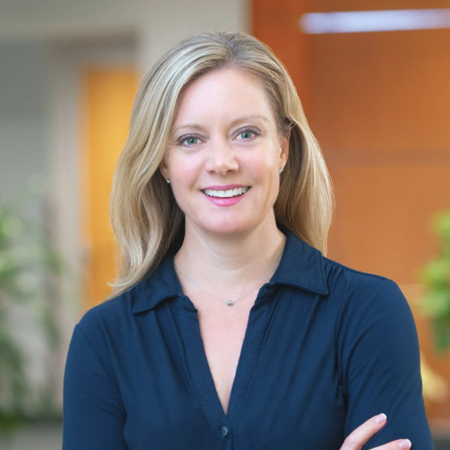Smiling woman with long blonde hair in a navy button-down shirt in a sunlit office
