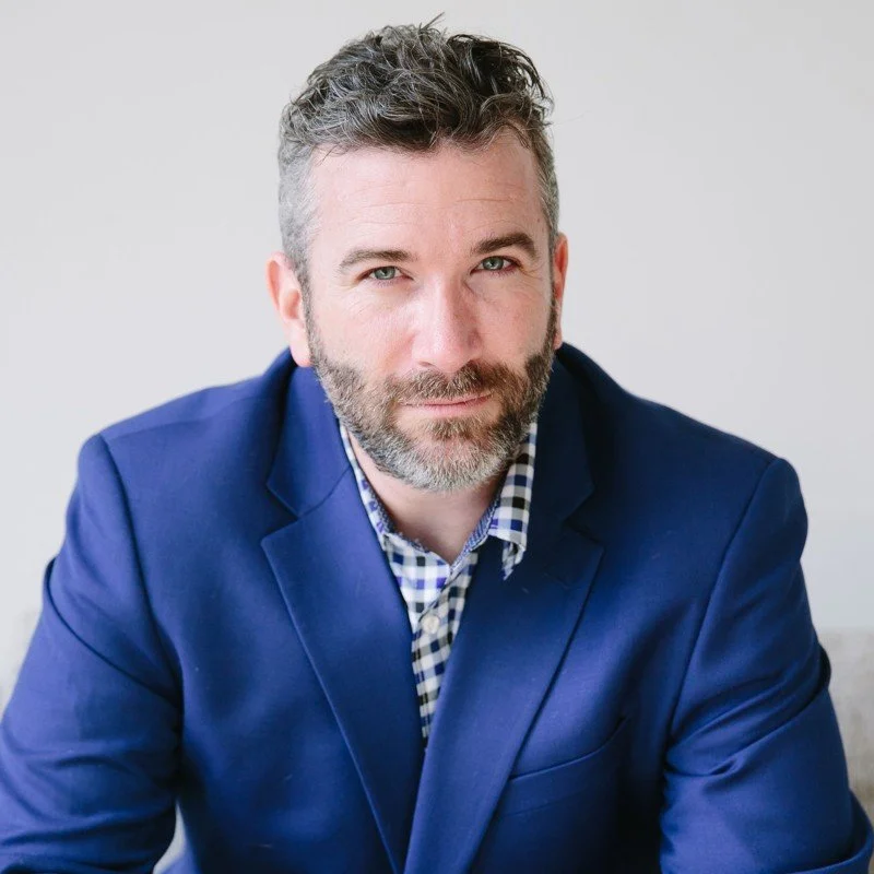 Man with grey hair and a beard in a bright blue blazer over a navy checked shirt